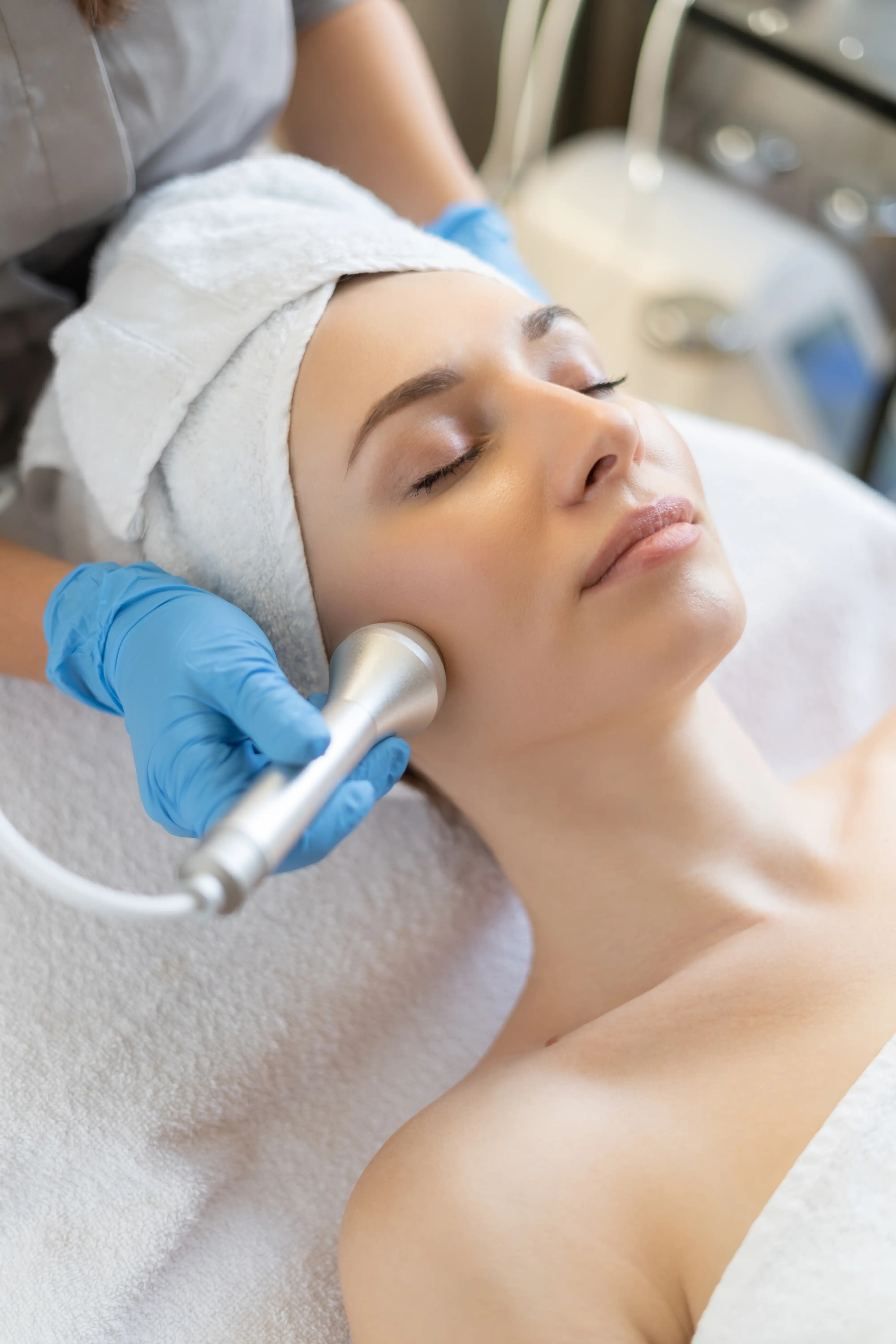 A gloved hand holding a metal handpiece to a reclining client’s cheek.